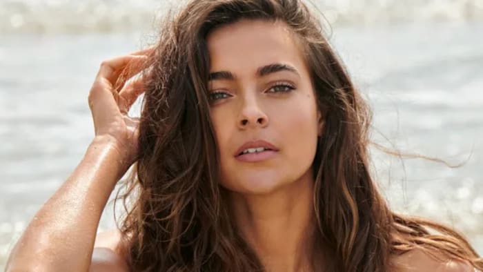 Ally Courtnall poses in front of the ocean water with her hand resting on the side of her head.